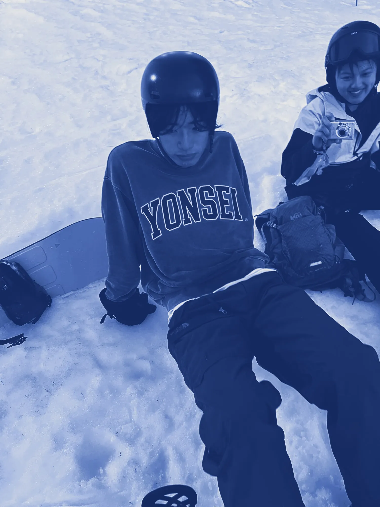 Ian sitting in the snow in a helmet beside his snowboard, a friend with a camera next to him, printed in cyanotype blue.
